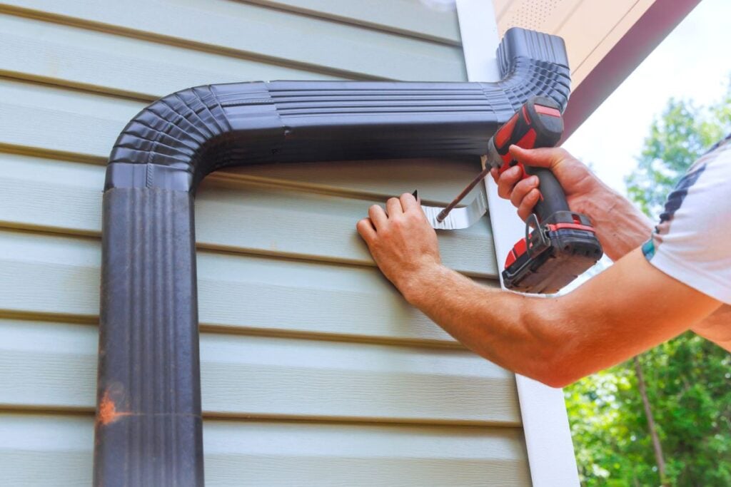 Worker installing rain gutters on roof, fitting downpipes, installing downspout bends